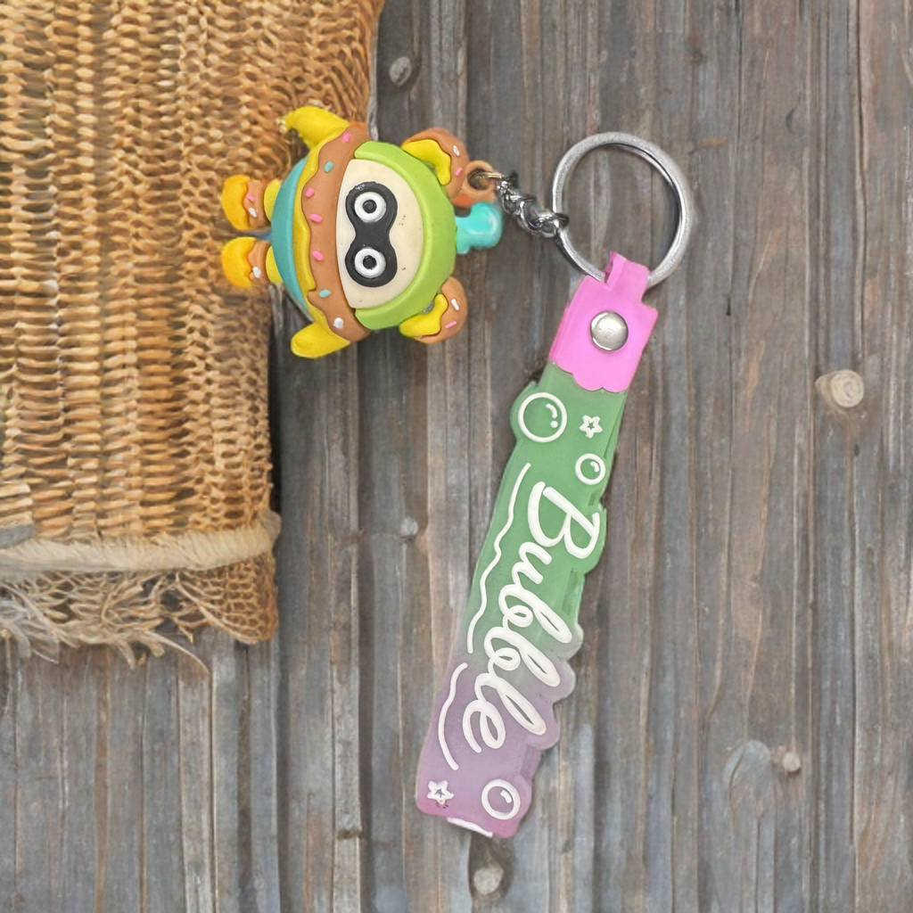 A colorful character keychain with a soft, plush design attached to a metal keyring. The keychain is displayed alongside a strap, possibly for scale.